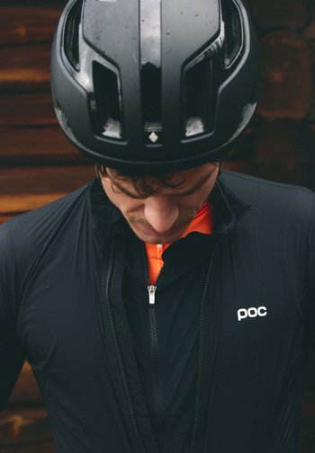 Mann som har på seg en svart sykkelhjelm og svart POC-jakke over en oransje skjorte, ser nedover mot en trebakgrunn.