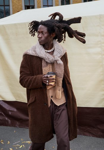 Persoon met lange dreadlocks, gekleed in gelaagde bruine kleding en een dikke sjaal, die een takeaway koffiebeker vasthoudt buiten.