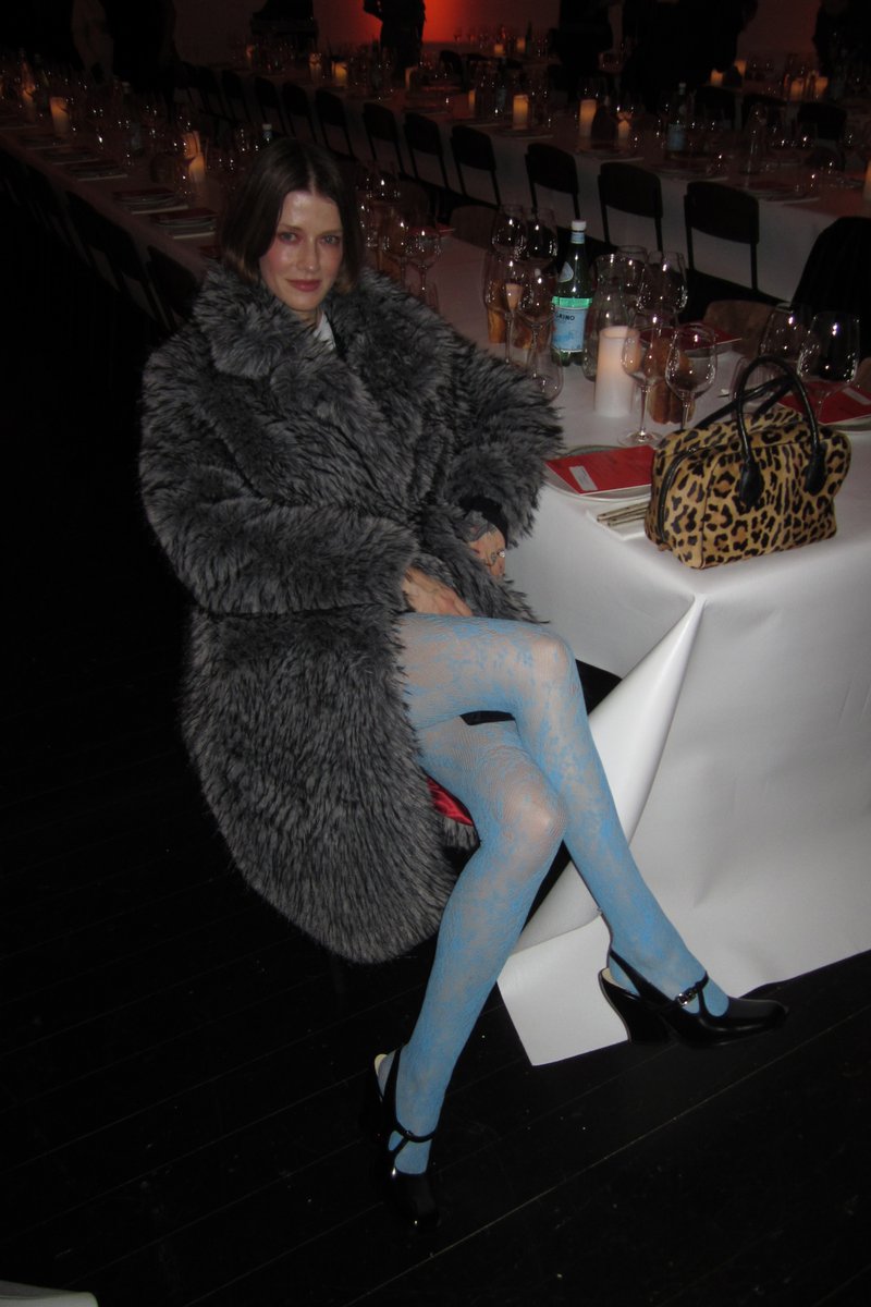 Eine Frau in einem grauen Pelzmantel sitzt elegant, trägt blau gemusterte Strumpfhosen und schwarze High Heels, während eine Handtasche im Leopardenmuster in der Nähe steht.