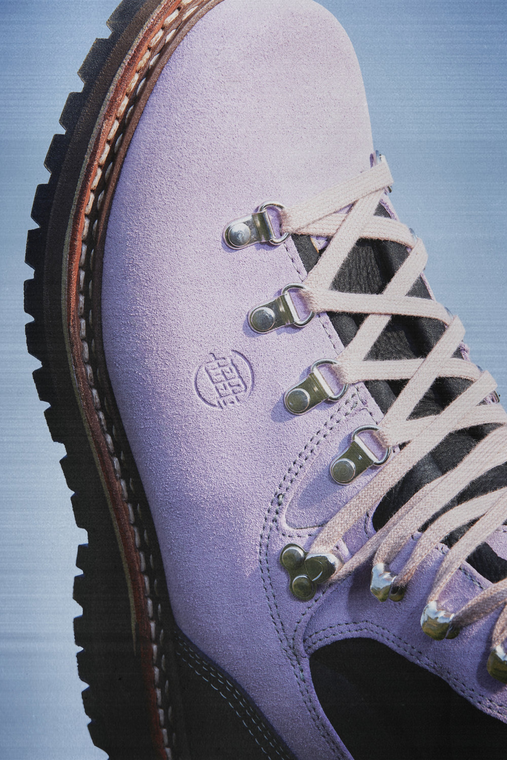 Close-up of a purple hiking boot with metal eyelets and laces, set against a light blue background. The boot features a textured surface and a sturdy sole.