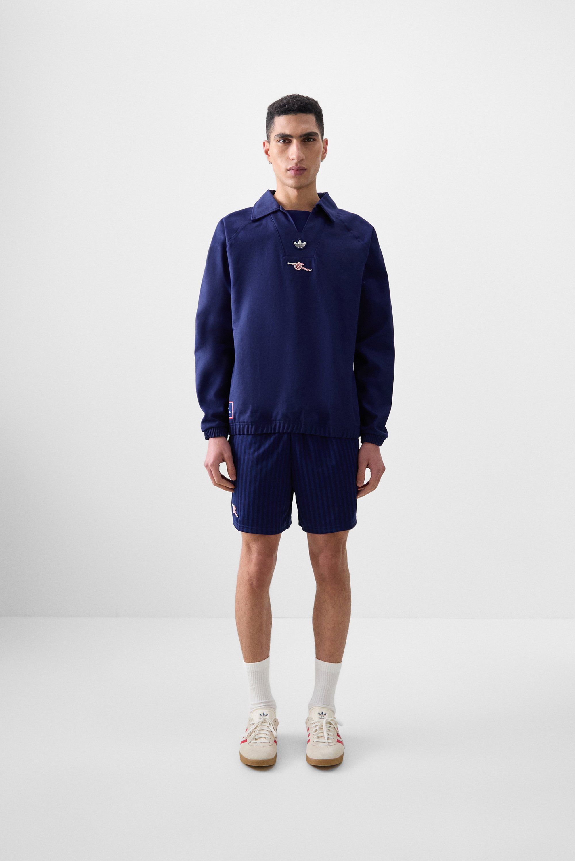 Een jonge man staat in een minimalistische omgeving, gekleed in een marineblauwe sweatshirt, gestreepte shorts en sneakers, met een zelfverzekerde uitdrukking.