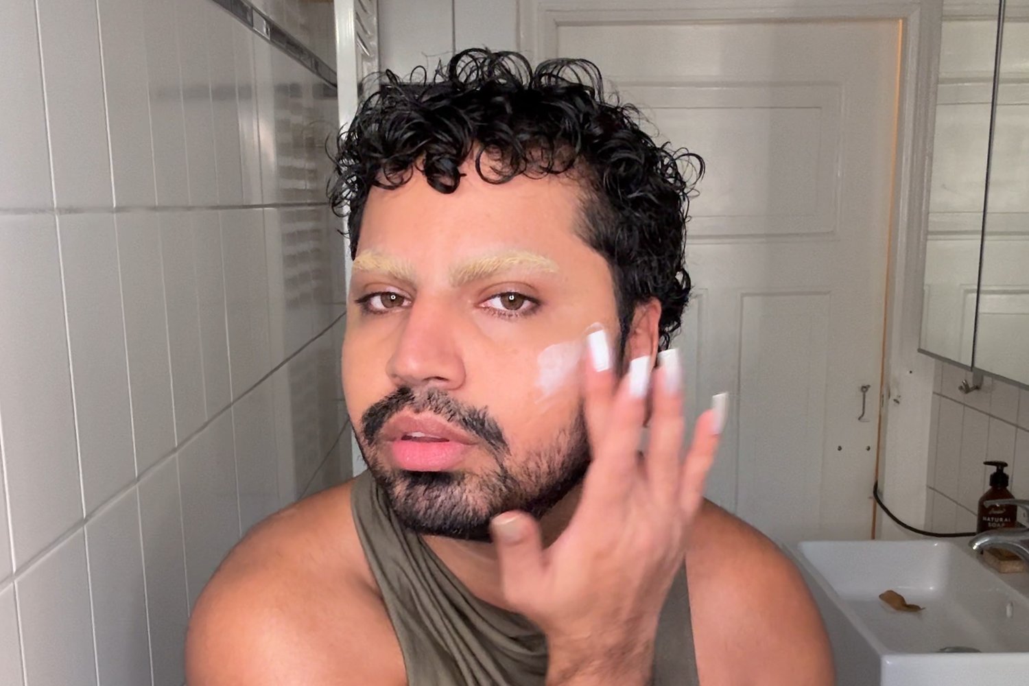 Persona con cabello rizado y cejas estilizadas aplicándose un producto para el cuidado de la piel en la cara, de pie en un baño bien iluminado con un espejo.