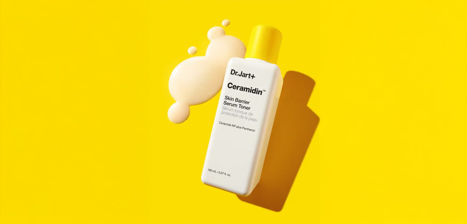 Weiße Flasche mit gelbem Verschluss, beschriftet mit "Dr.Jart+ Ceramidin Skin Barrier Serum Toner", daneben ein cremefarbener Tropfen vor gelbem Hintergrund.