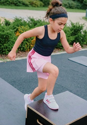 Junges Mädchen in Sportkleidung steigt im Freien auf eine Trainingsbox, konzentriert auf die Fitnessaktivität mit Grünpflanzen im Hintergrund.