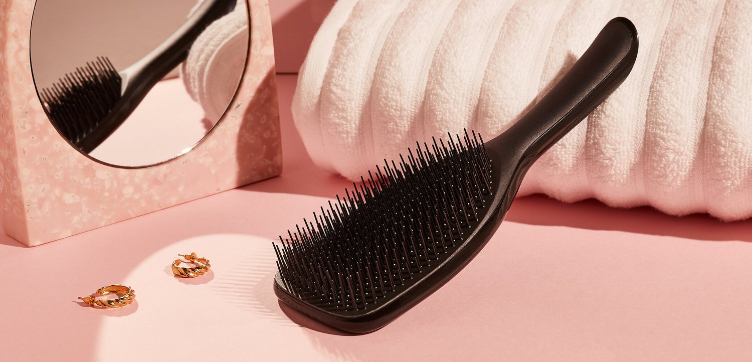 Brosse à cheveux plate noire, boucles d'oreilles anneaux dorés, serviette blanche et un petit miroir rond sur une surface rose douce.