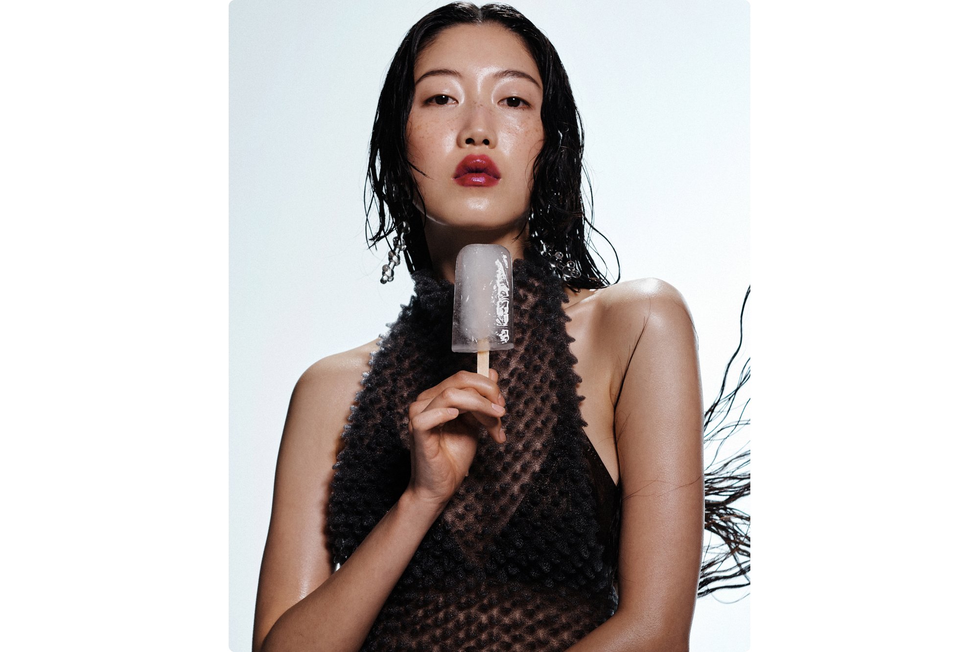A young woman with wet, tousled hair holds a popsicle, wearing a sheer, textured black outfit against a light background.