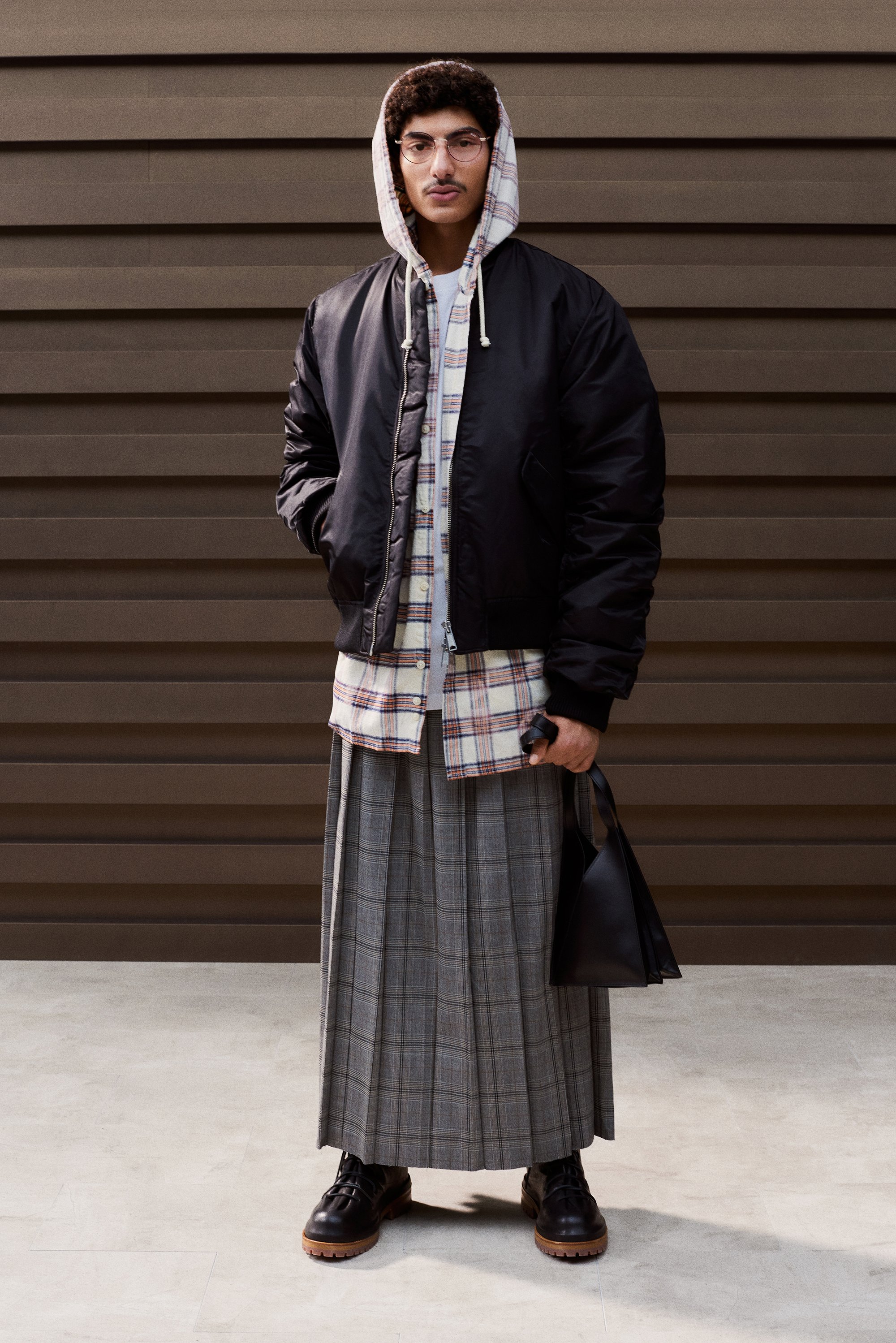Person stands against a textured brown wall, wearing a black bomber jacket over a plaid shirt and gray pleated skirt, holding a black bag.