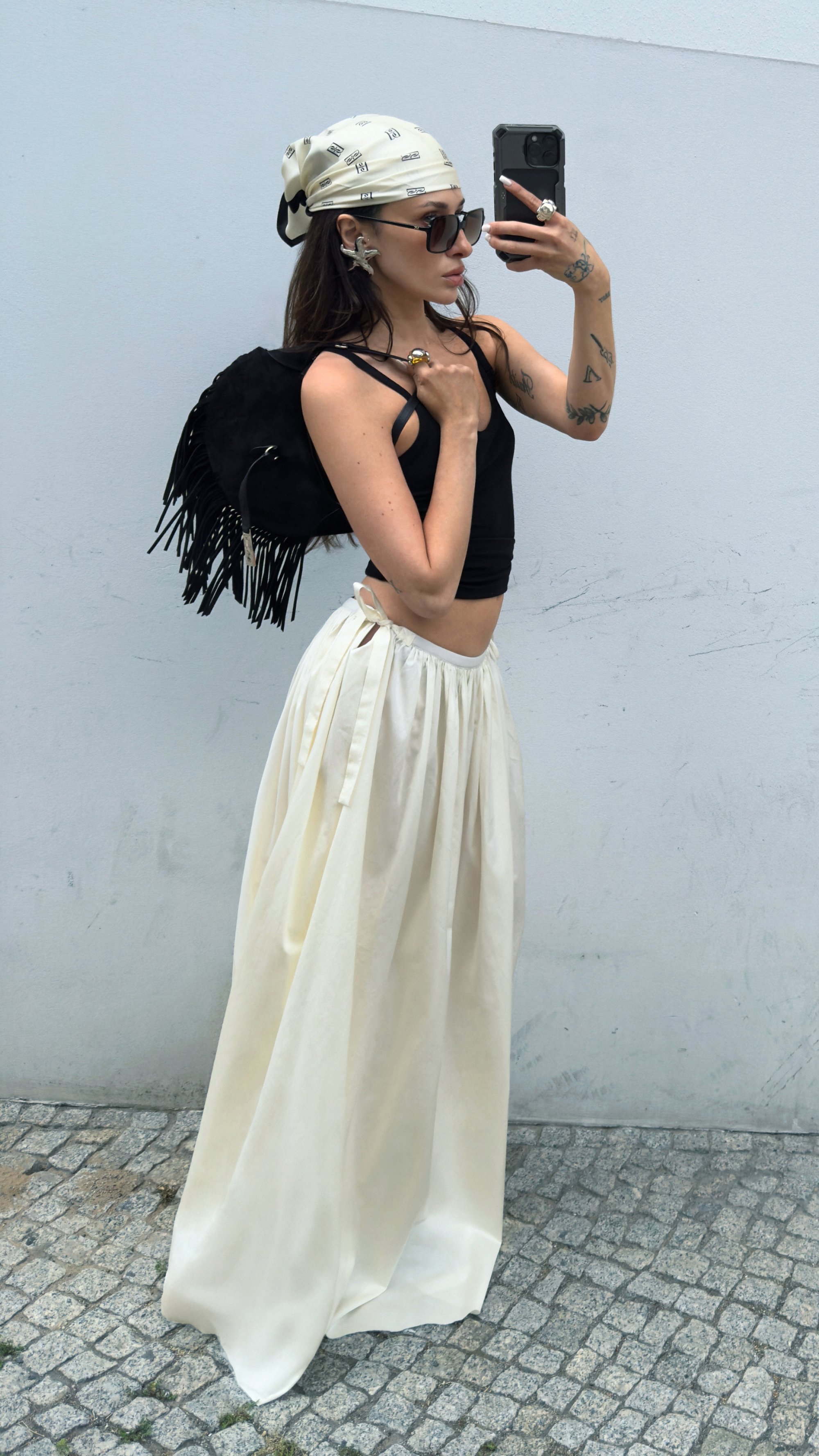 Zaino in suede nero con frange, crop top nero e gonna bianca fluida con laccetti laterali. Gli accessori includono occhiali da sole e sciarpa stile fantasia.