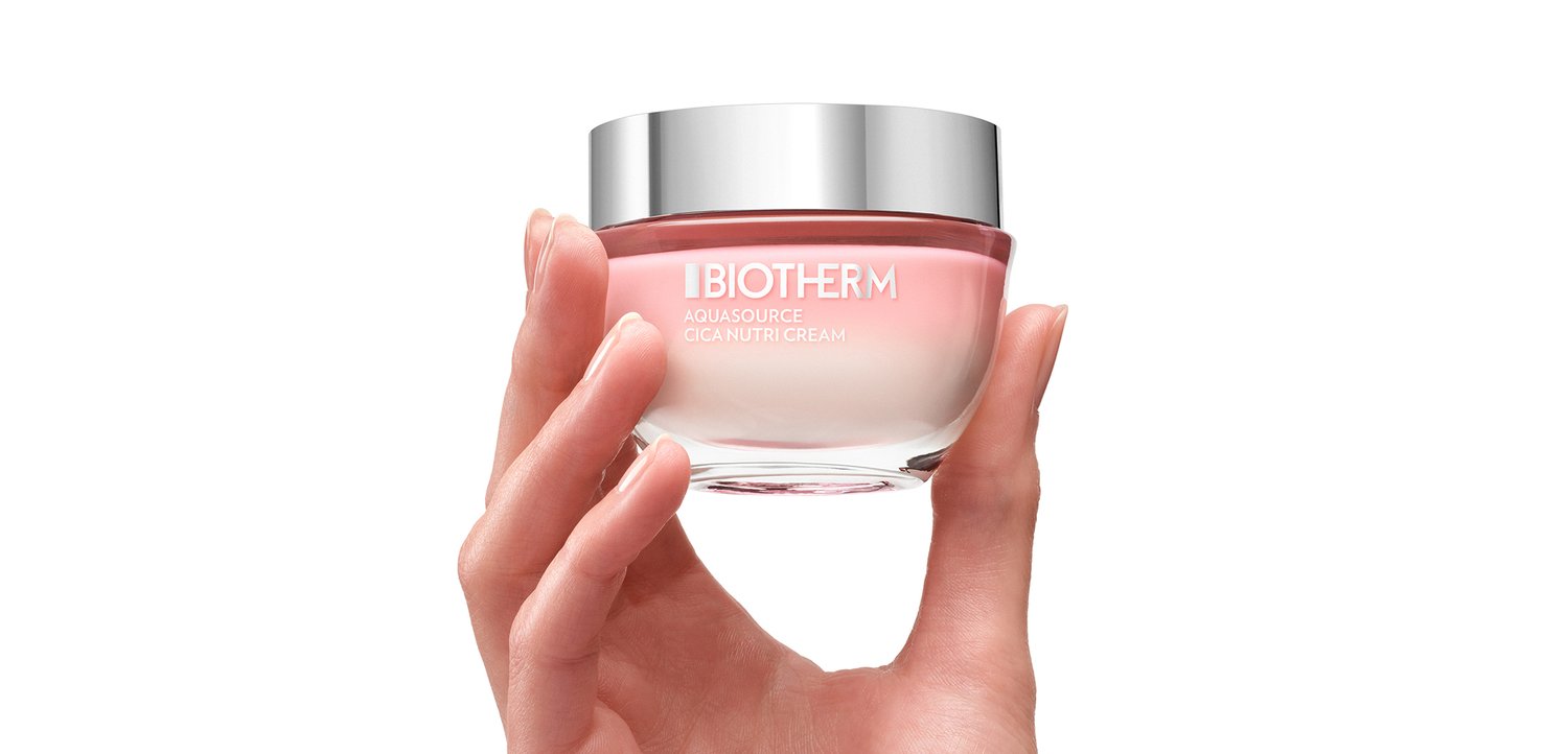 Een hand houdt een glazen pot van Biotherm Aquasource Cica Nutri Cream vast, met een zilveren deksel en een roze verloopcrème erin, tegen een witte achtergrond.