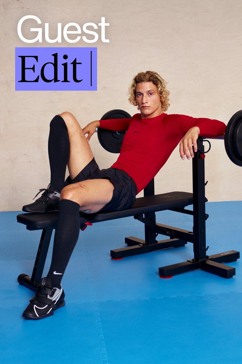 Een persoon met krullend haar ligt op een gewichtbank, gekleed in een rood langshirt, zwarte shorts en kniesocks in een sportschoolsetting.