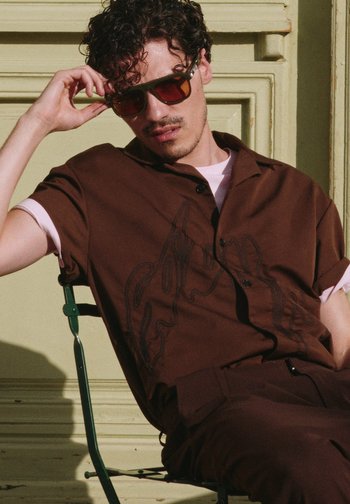 Άνδρας με σγουρά μαλλιά που φοράει καφέ πουκάμισο και παντελόνι, κάθεται σε πράσινη μεταλλική καρέκλα, προσαρμόζοντας μεγάλα καφέ γυαλιά ηλίου.