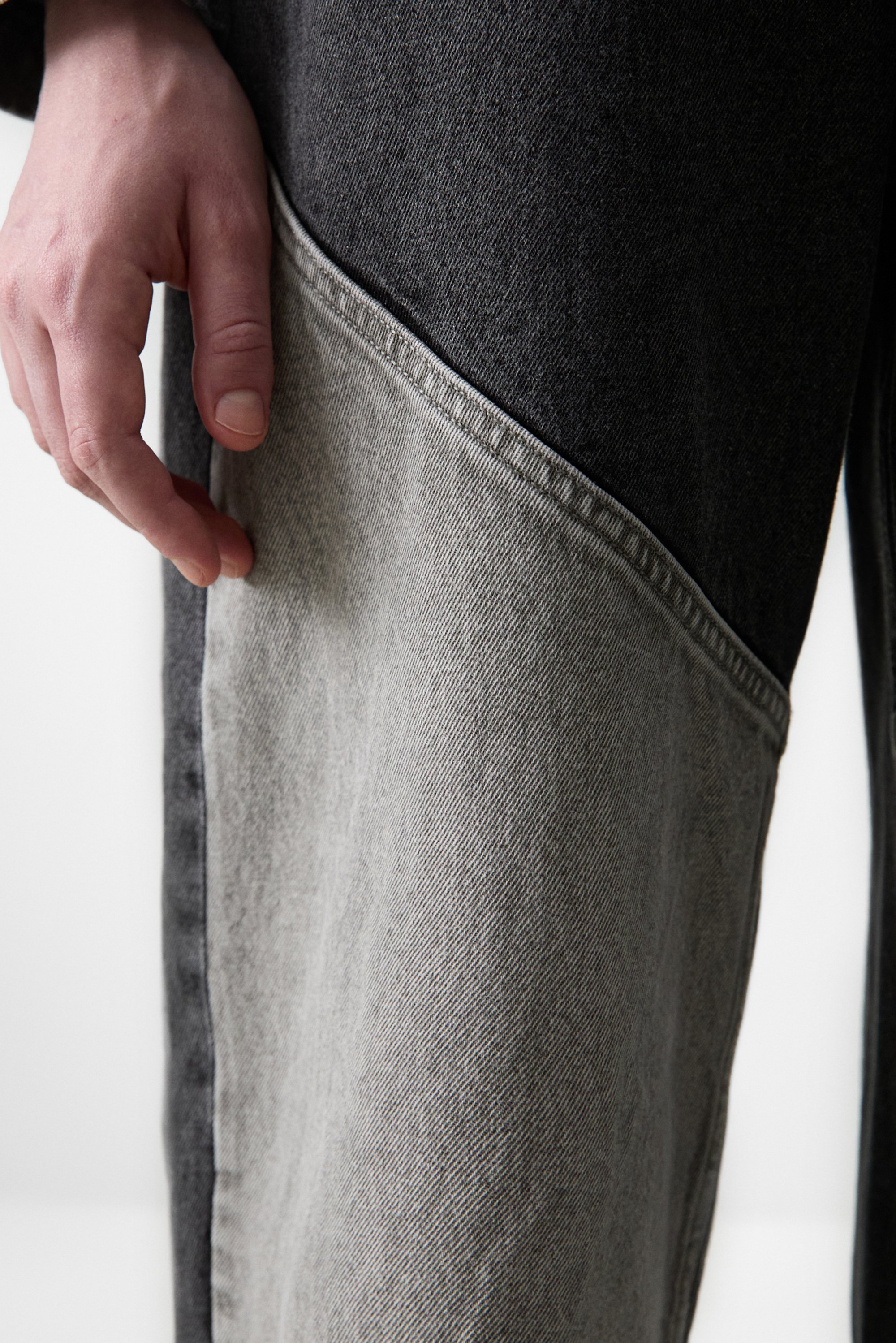 Un primo piano di una mano che tocca un capo d'abbigliamento con un design a blocchi di colore, caratterizzato da sezioni di denim scuro e chiaro. Lo sfondo è neutro.
