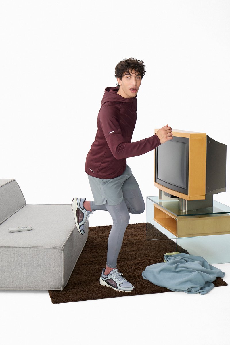 Young man in a maroon hoodie sitting and lying on a modern gray couch, with a colorful blanket, interacting with an old television.