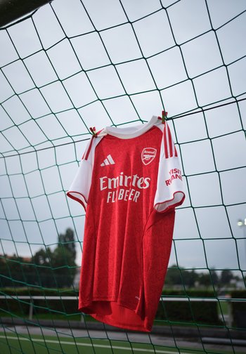 Punase ja valge Arsenal'i jalgpallisärk, mis ripub rohelisel värava võrgul õues, taustal on pilvine taevas.