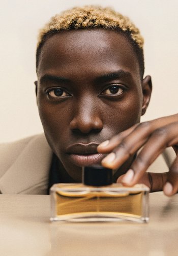 Flacon de parfum avec une base transparente et un liquide doré, surmonté d'un bouchon noir. L'arrière-plan présente une couleur neutre avec des textures douces.