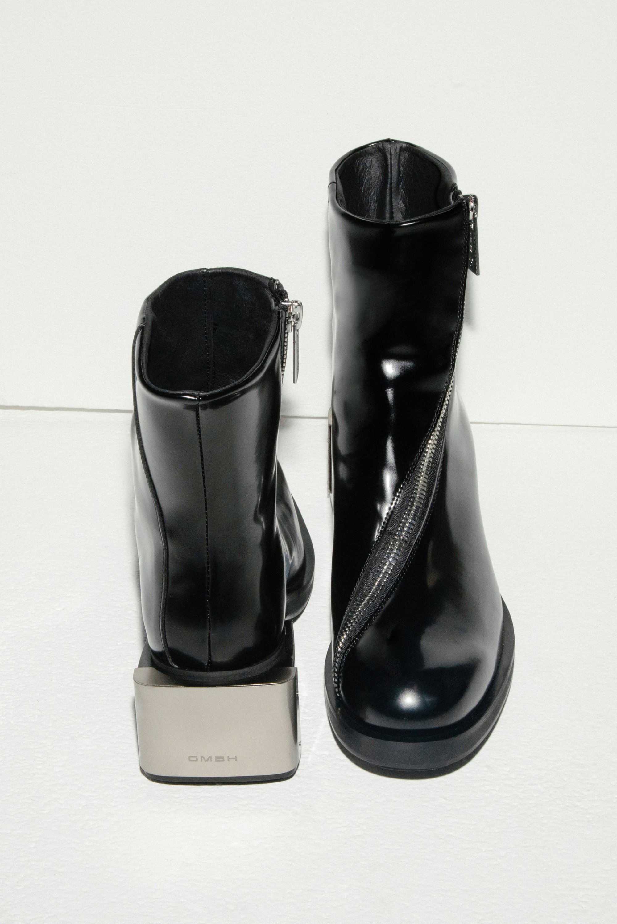 Schwarze Leder-Stiefeletten mit einem Seitenreißverschluss und einem metallischen Blockabsatz, vor einem weißen Hintergrund präsentiert.