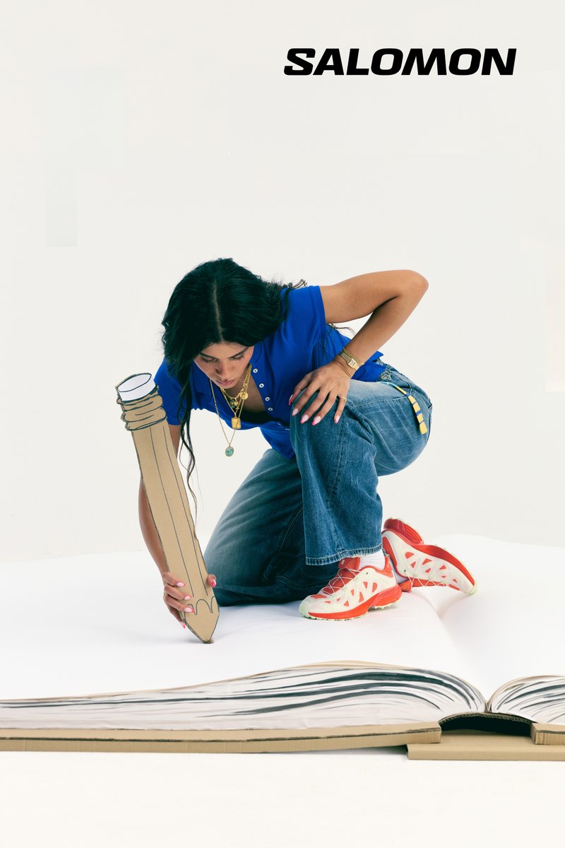 Una mujer con una camisa azul y vaqueros se arrodilla sobre un libro grande sosteniendo un lápiz de cartón grande, vistiendo zapatillas rojas y blancas, con el logo de Salomon arriba.