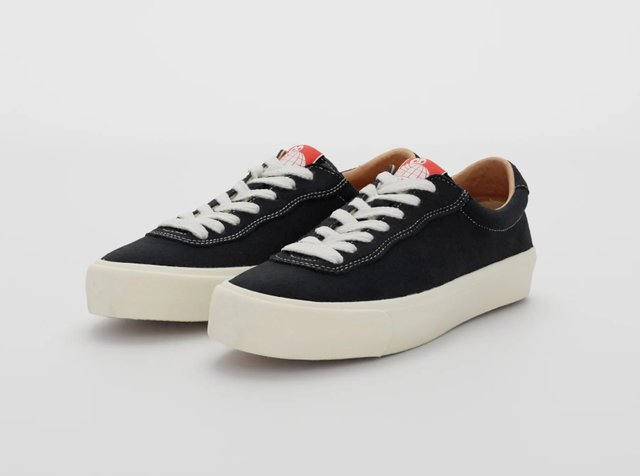 Sorte kanvas sneakers med hvite lisser, kremfargede gummisåler og en rød detalj ved hælen. Rund form med hvite sømdetaljer.