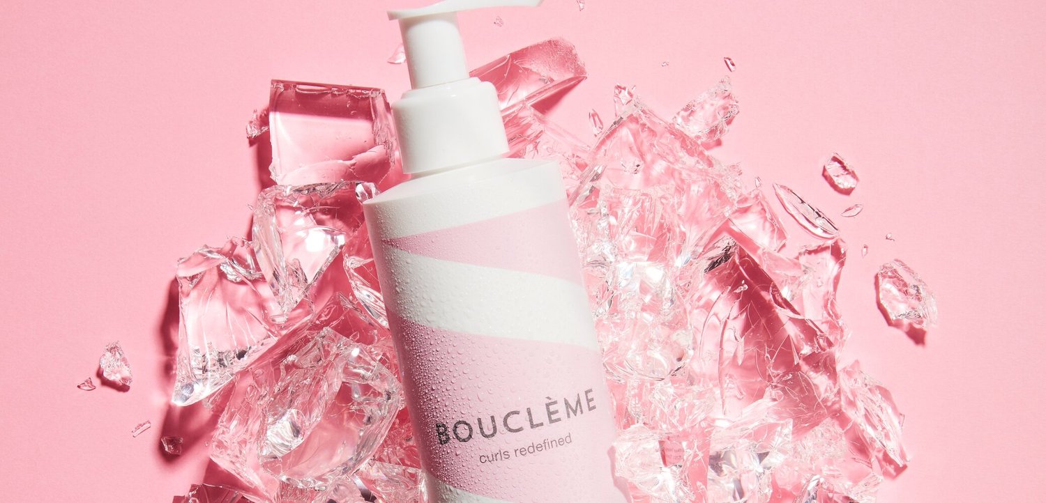 Weisse und rosa Bouclème-Lotionsflasche, umgeben von klaren Eiswürfeln auf einem einfarbigen pinken Hintergrund.