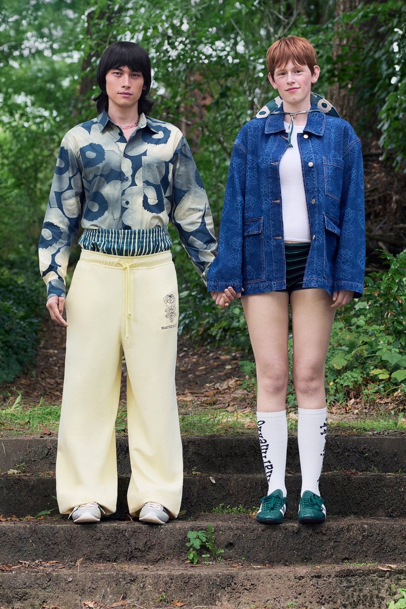 Two individuals stand side by side. One is dressed in a blue floral shirt and yellow wide trousers; the other is wearing a denim jacket and striped shorts.