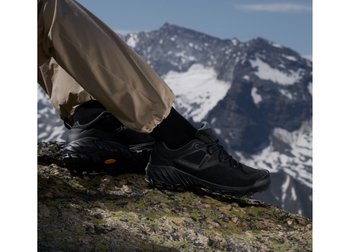 Eine Nahaufnahme von schwarzen Wanderschuhen auf felsigem Terrain, mit schneebedeckten Bergen im Hintergrund und einem bewölkten Himmel darüber.