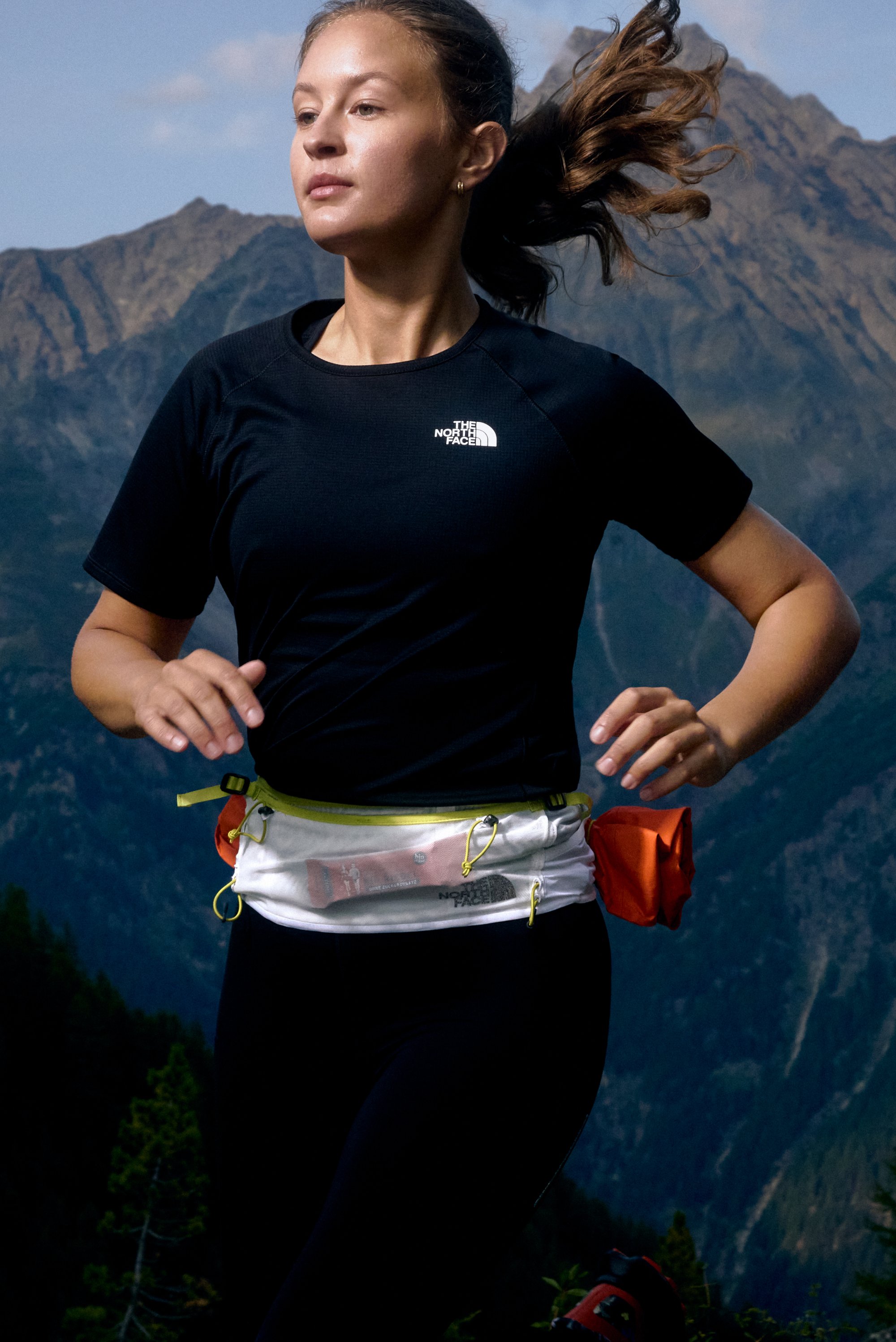 Camiseta atlética negra y leggings usados con una riñonera blanca ligera con acentos en amarillo y naranja, en un fondo de montaña.