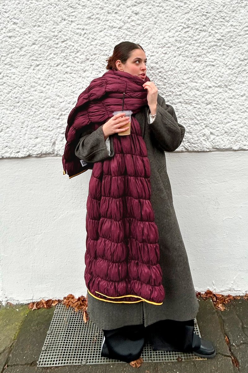 Bourgognefarvet polstret tørklæde med quiltet tekstur og blank finish. Båret over en grå frakke, holdt af en person med en drink.