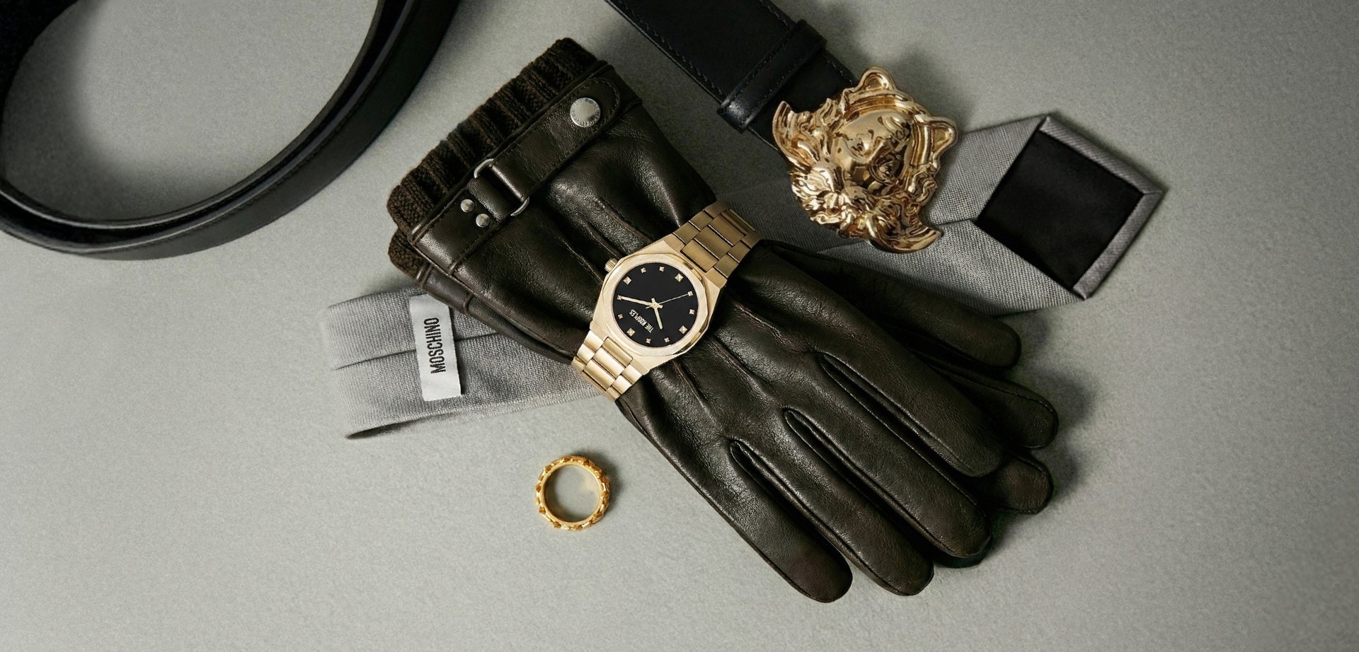 Black leather gloves featuring brown knitted accents, a gold watch with a black dial, a gold ring, and a black belt with an ornate buckle.