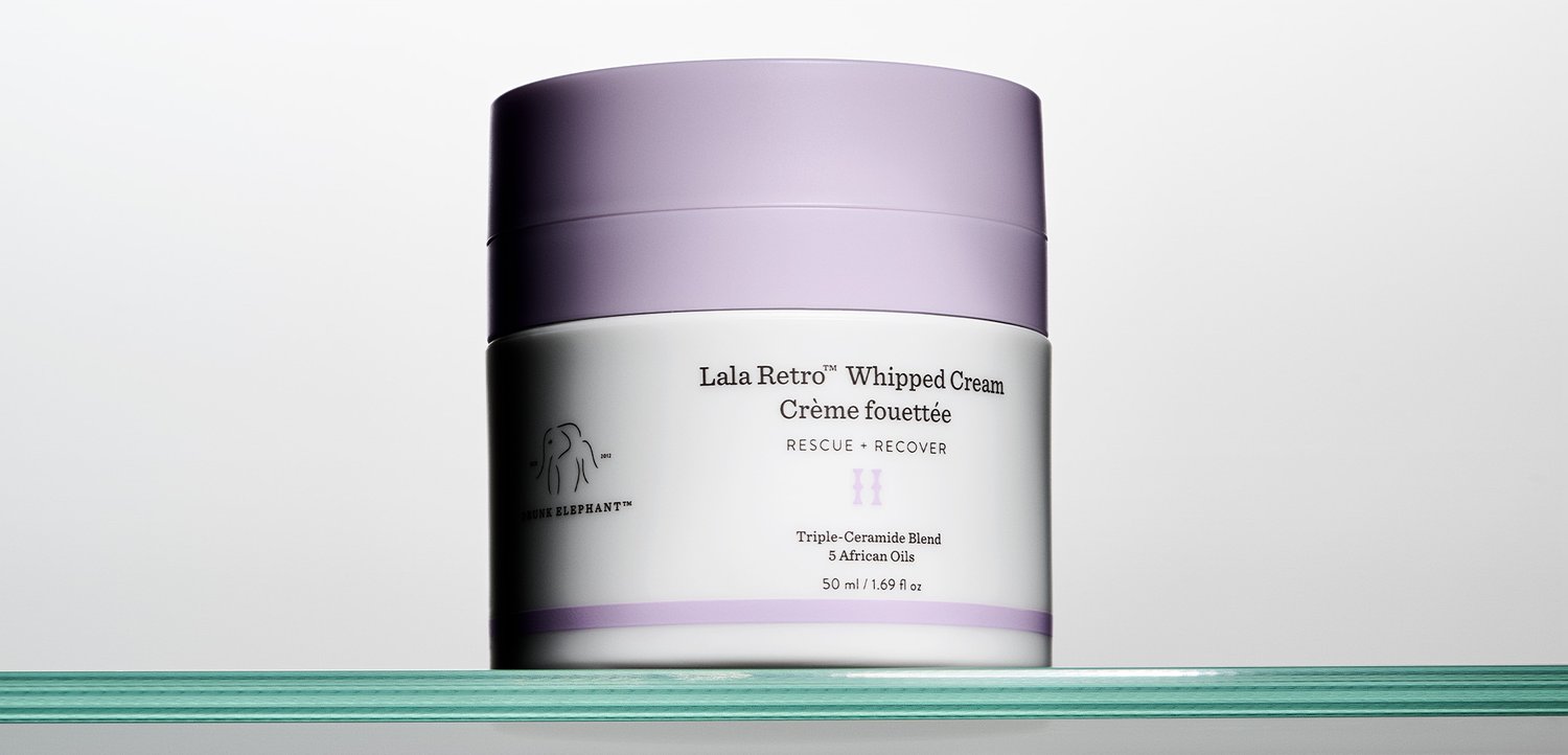 Glas mit Drunk Elephant Lala Retro Whipped Cream mit lavendelfarbenem Deckel und weißem Etikett, das auf einem klaren Glasregal vor einem schlichten Hintergrund steht.