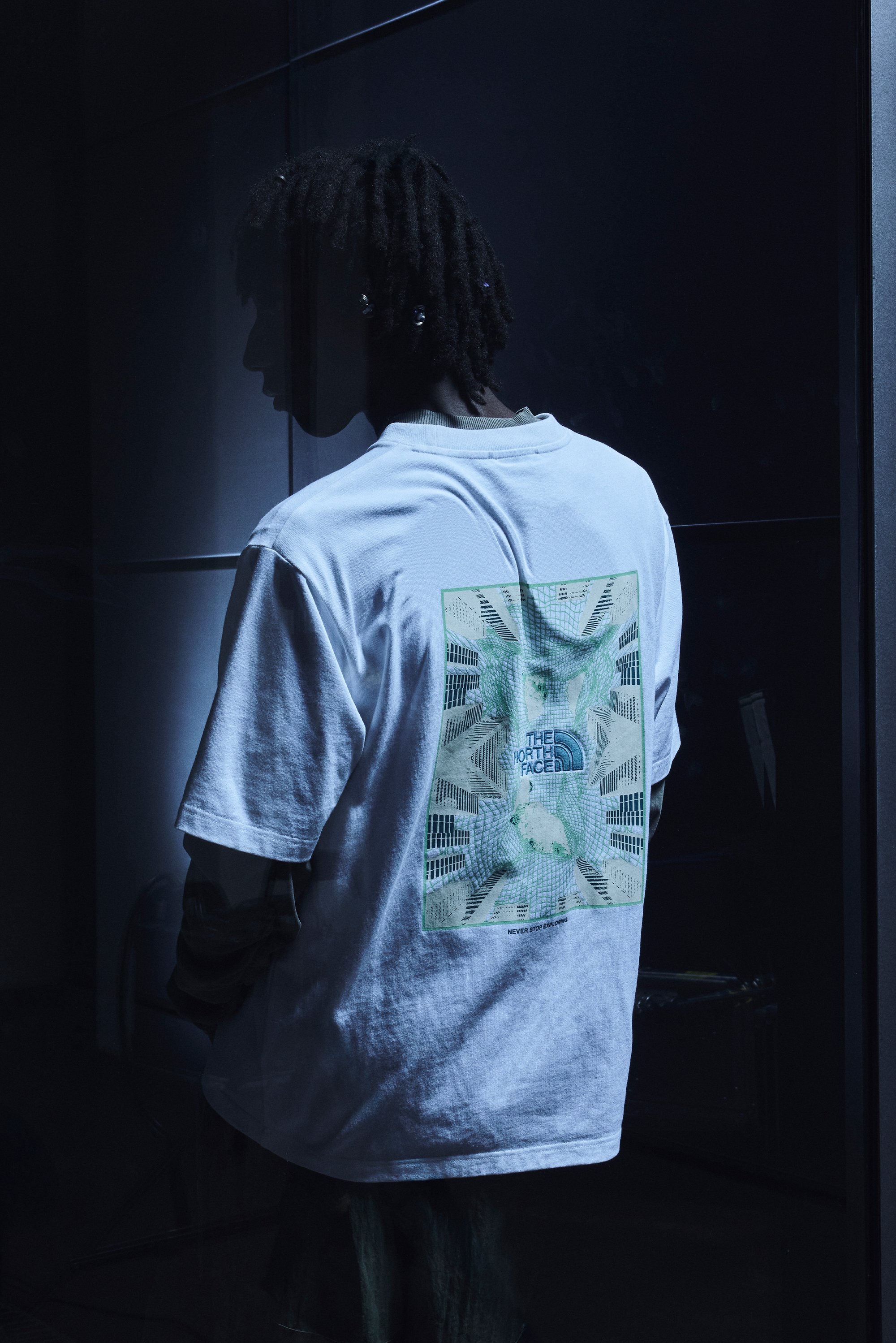 Una persona con dreadlocks indossa una T-shirt bianca con un design grafico di The North Face, su uno sfondo scuro.