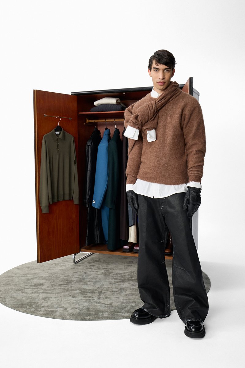 A man sits on the floor beside an open wardrobe filled with various clothing. He wears a brown sweater and gloves, looking at the camera.