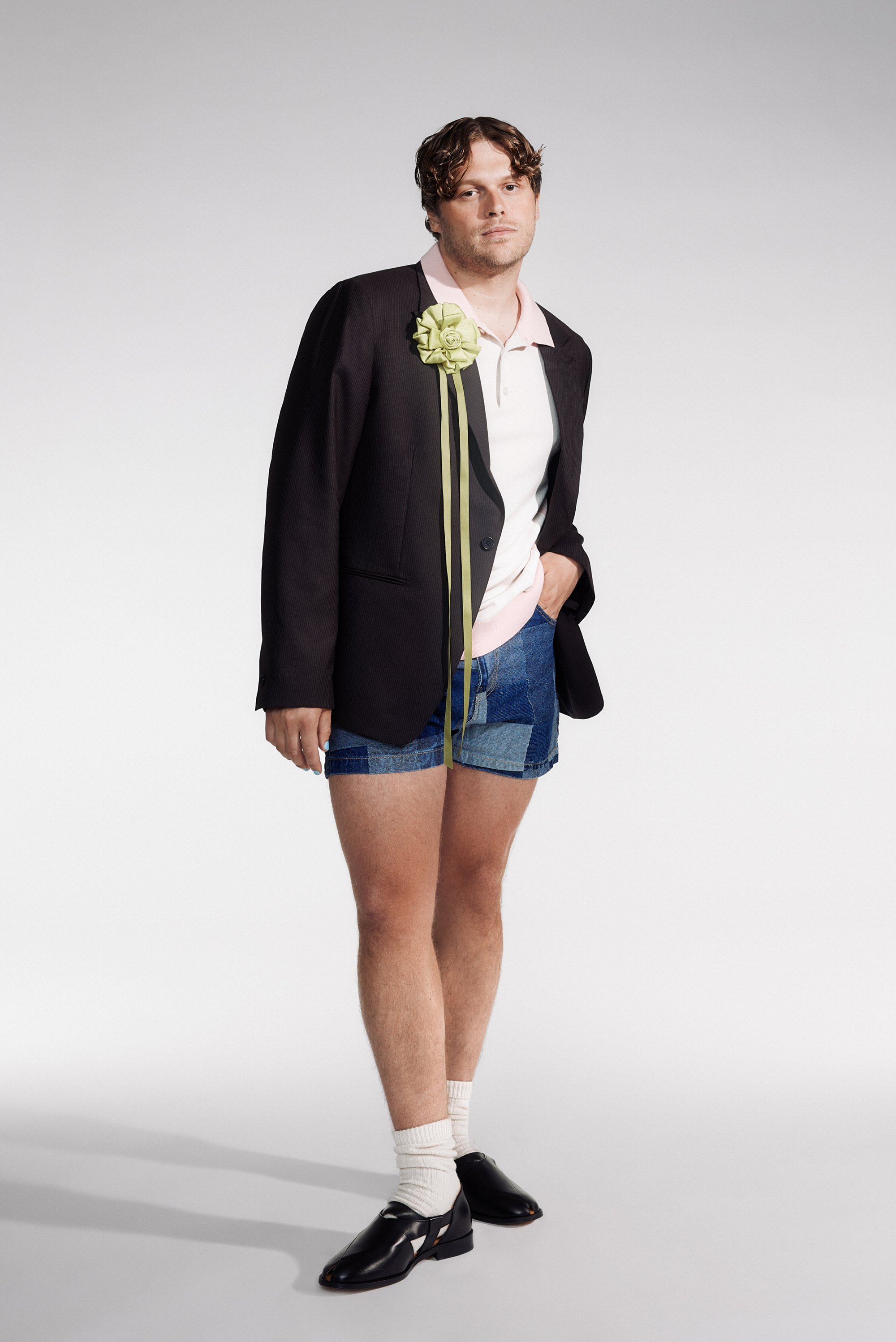 Una persona se encuentra en una pose relajada, vistiendo un blazer negro sobre una camiseta tipo polo, pantalones cortos de mezclilla y zapatos negros, con una flor decorativa en el blazer.