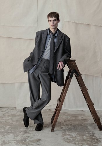 Jonge man in een loszittend grijs pak met das leunt tegen een houten ladder voor een gestructureerde beige achtergrond op een betonnen vloer.