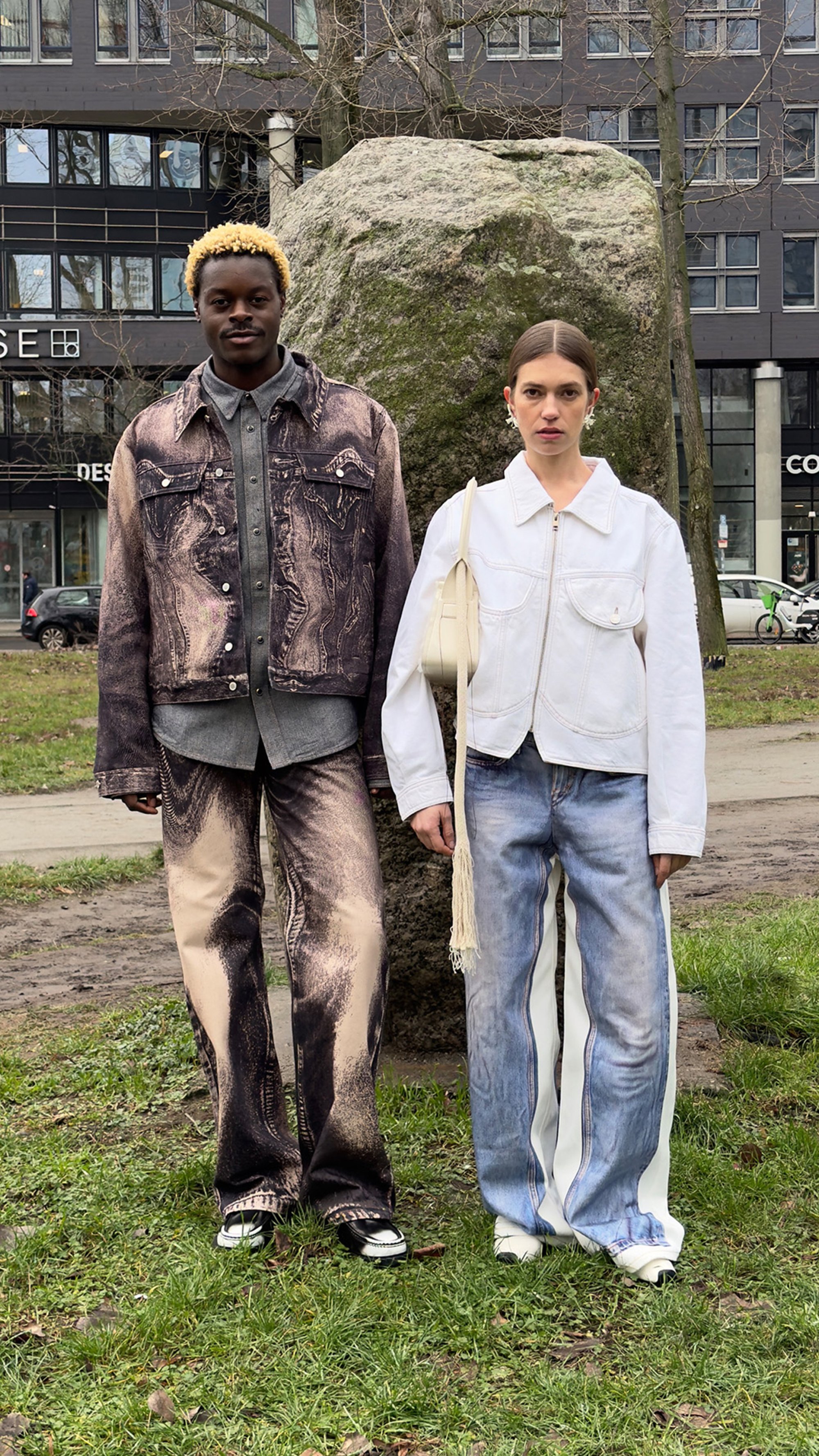 Dos modelos están de pie junto a una gran roca en una zona grassy, mostrando atuendos modernos: una con un conjunto de mezclilla estampada y la otra con una chaqueta blanca.