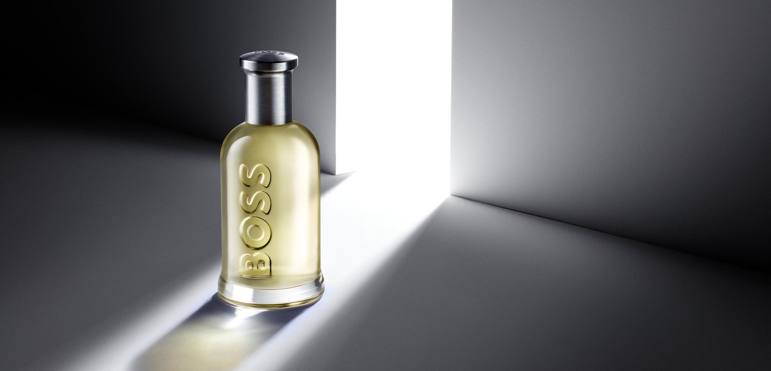 Una bottiglia di profumo traslucida etichettata BOSS si trova su una superficie con una luce intensa che attraversa una porta aperta sullo sfondo.