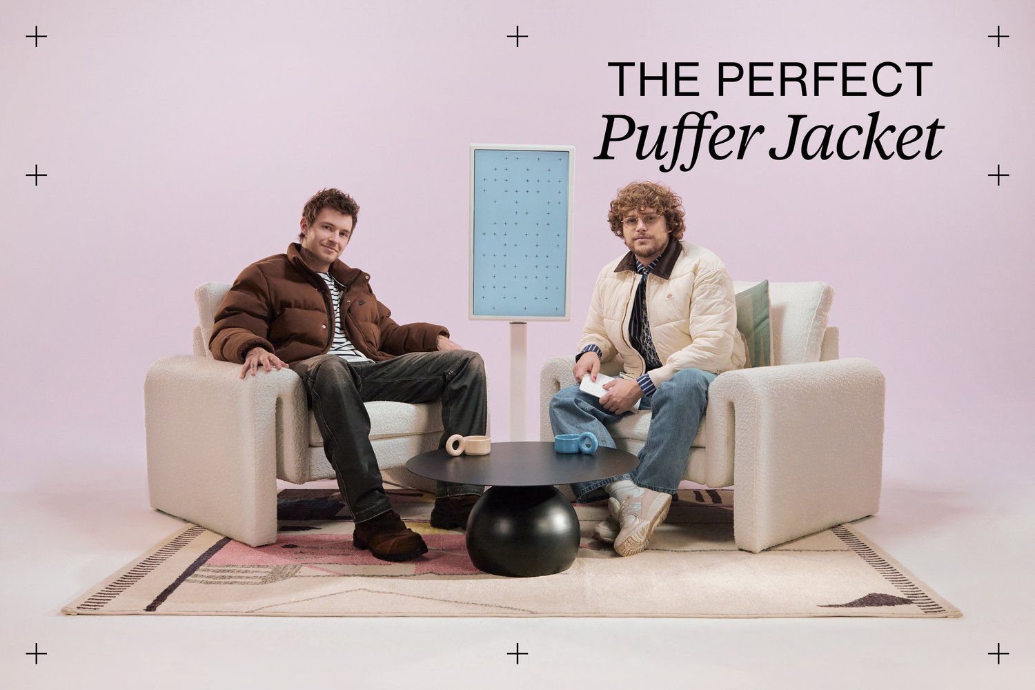 Two men sit on cream sofas in a pastel pink setting. A black table holds colorful mugs while a light blue display stands between them.