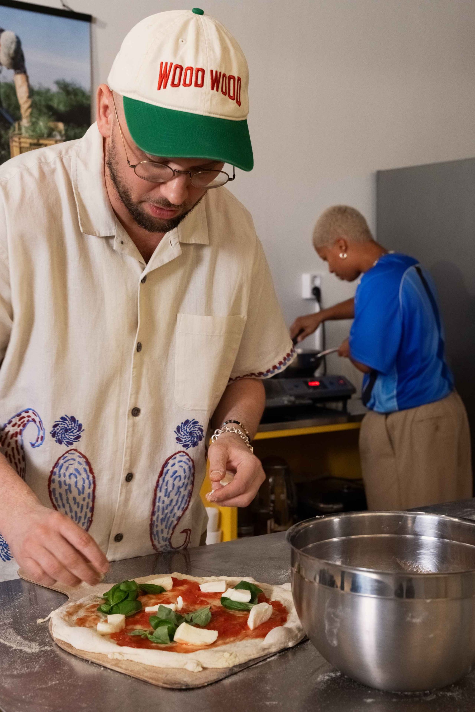 Witte pizzadeeg belegd met tomatensaus, mozzarella en verse spinazie. Een man in een beige shirt voegt ingrediënten toe, met een roestvrijstalen kom in de buurt.