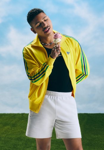 Mann, der eine gelbe Jacke und weiße Shorts trägt, leckt im Freien an einem Eis mit zwei Kugeln vor blauem Himmel.