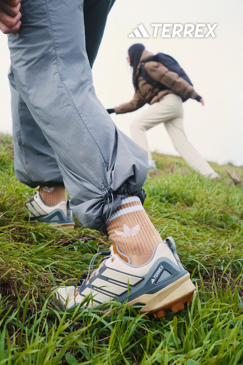 Terrex buitenschoenen met een crème en grijs ontwerp, voorzien van een stevige zool, vetersluiting en accentkleuren. Draagt beige sokken en grijze broek.