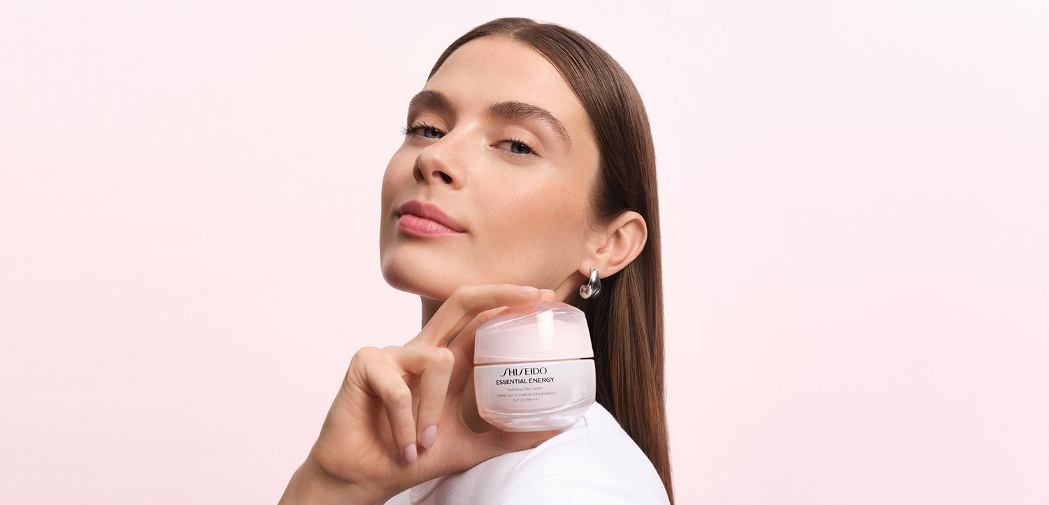 Kobieta z czystą cerą trzyma słoik kremu do pielęgnacji skóry Shiseido Essential Energy blisko twarzy, na miękkim różowym tle.