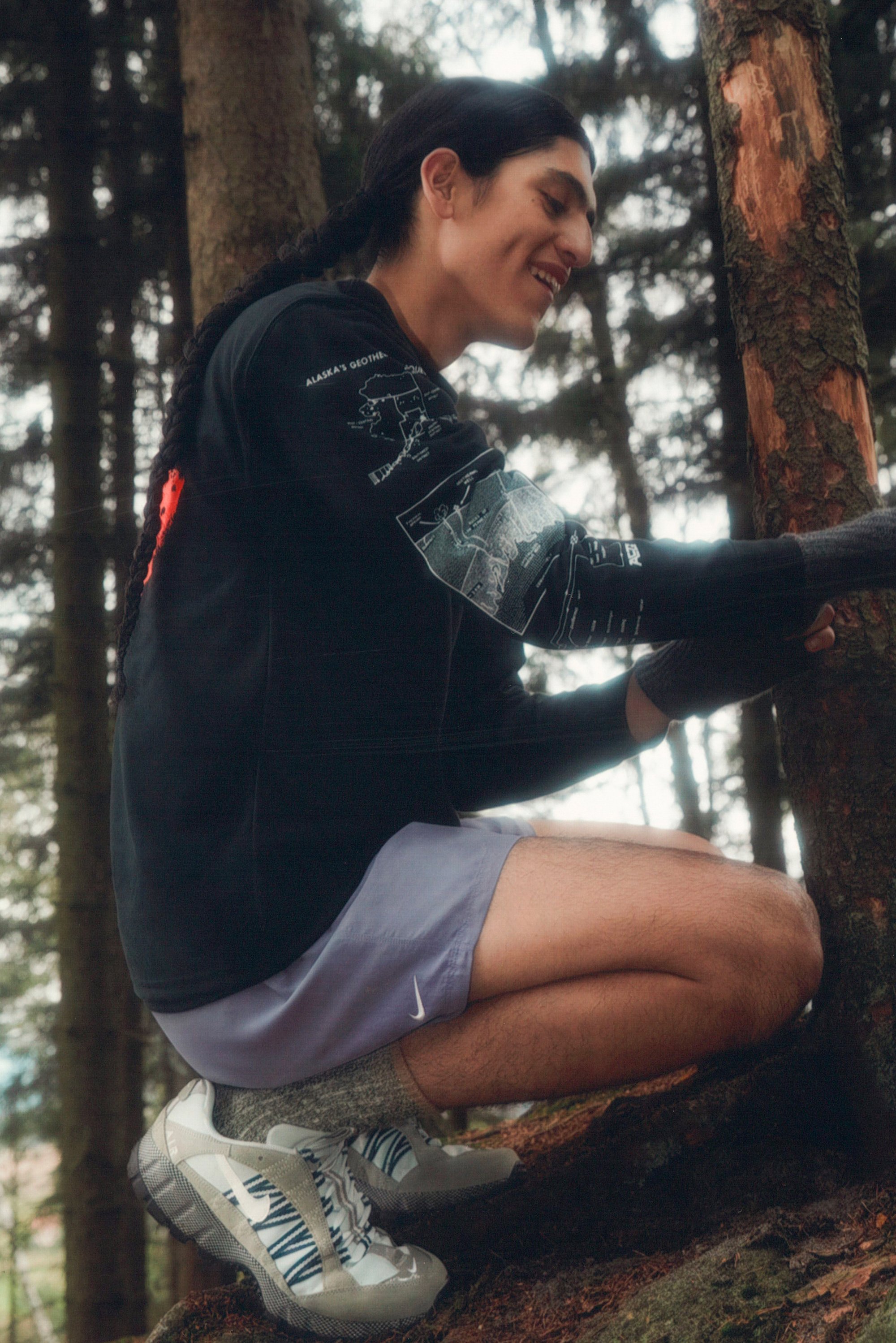 Un joven adulto con el pelo largo, que lleva una camiseta negra de manga larga y pantalones cortos morados, se arrodilla junto a un árbol en un entorno forestal.