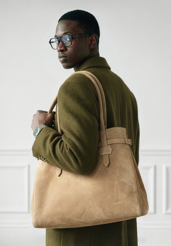 Άνδρας που φορά γυαλιά και παλτό ελιάς κρατά μια μεγάλη μπεζ τσάντα από σουέτ στον ώμο του, κοιτάζοντας πλάγια.