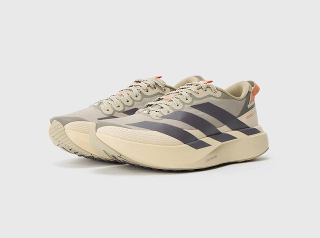 Par de zapatillas de running beige con suelas gruesas, diseño a rayas negras, lengüetas naranjas en el talón y cordones, sobre un fondo claro y liso.