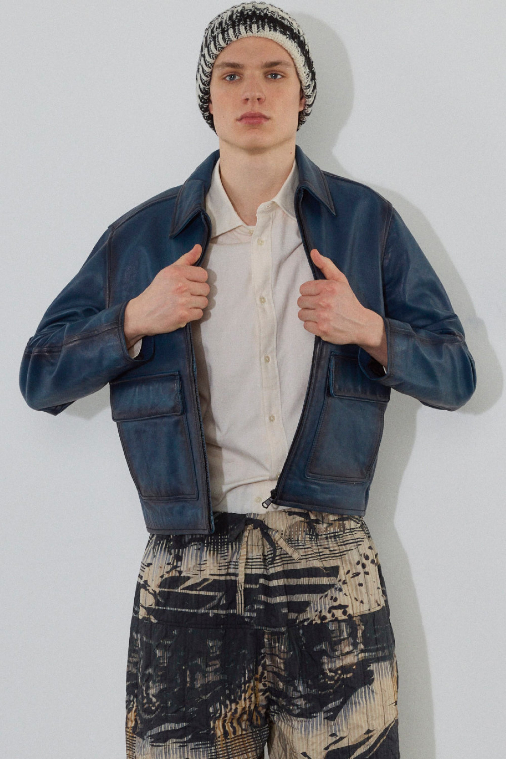 Un joven posa con una chaqueta de cuero azul sobre una camiseta blanca, combinada con pantalones cortos de estampado y un gorro de punto en blanco y negro.