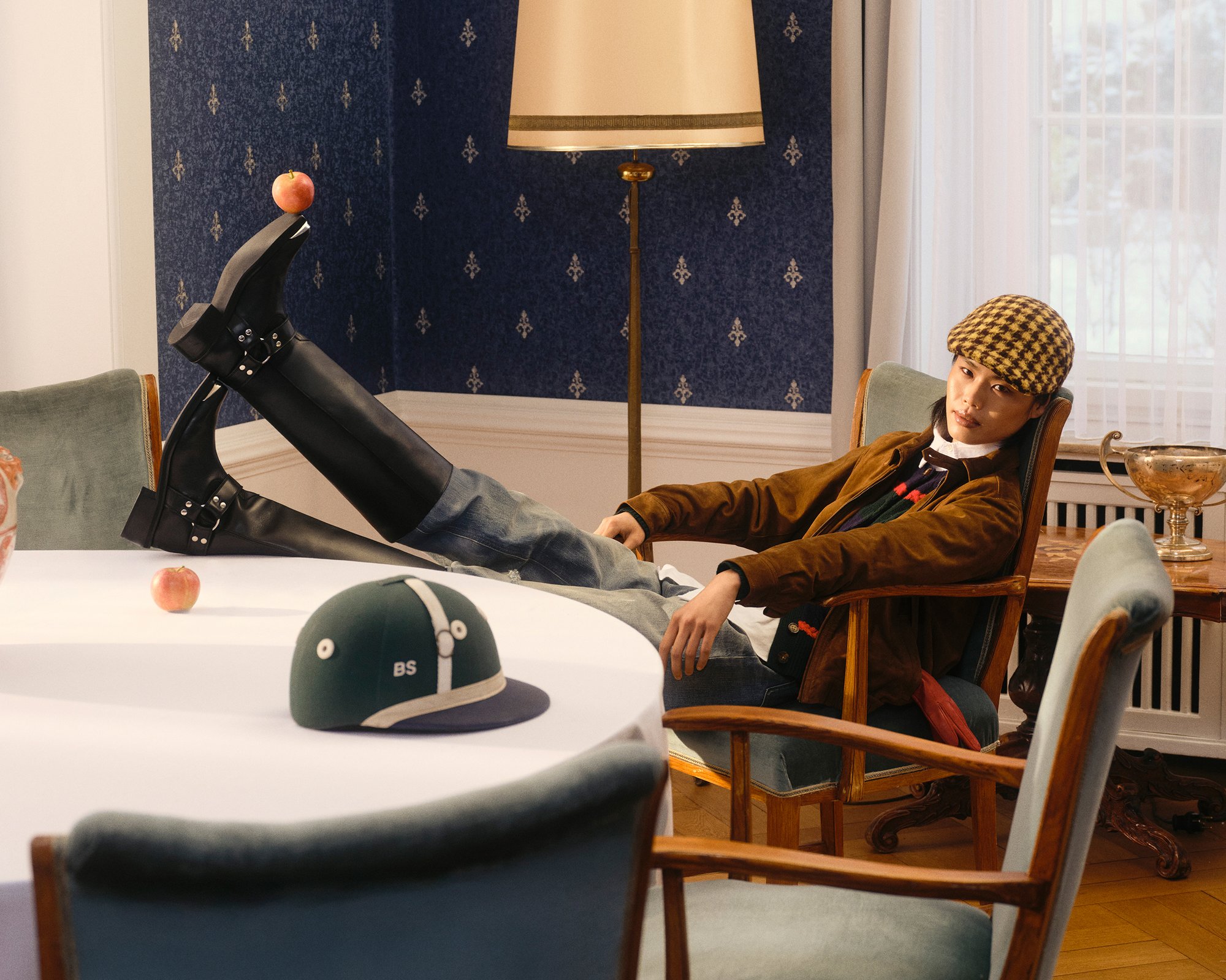 Una persona está sentada en una silla con las piernas cruzadas, vistiendo unas botas negras altas y una gorra a cuadros. Una manzana reposa sobre la mesa, junto a un sombrero verde.