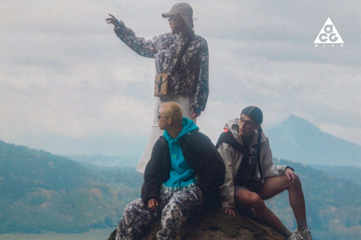 Tres jóvenes posan sobre una roca al aire libre. Uno señala mientras los otros dos están sentados, vestidos con ropa de exterior casual y a la moda, frente a un nebuloso fondo montañoso.
