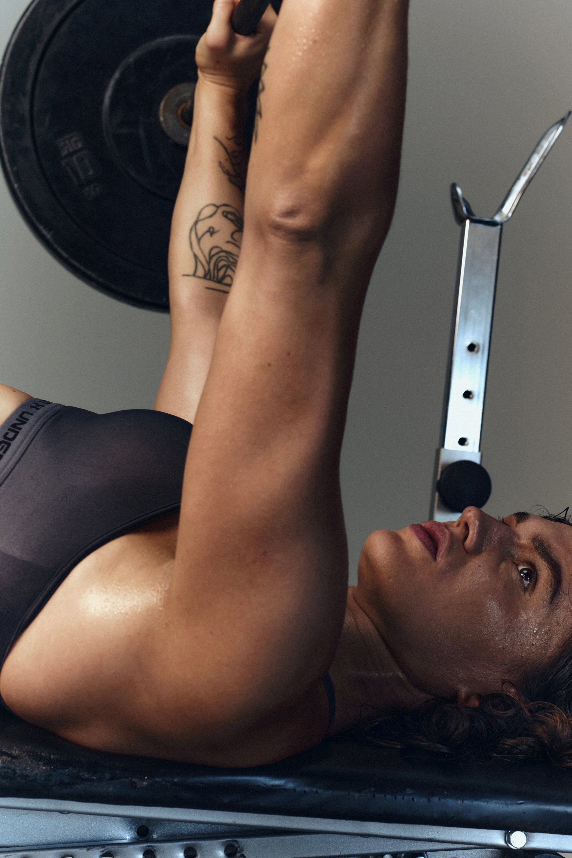 A muscular woman bench presses a barbell, focusing intensely. She has tattoos on her arm and sweat glistening on her skin.