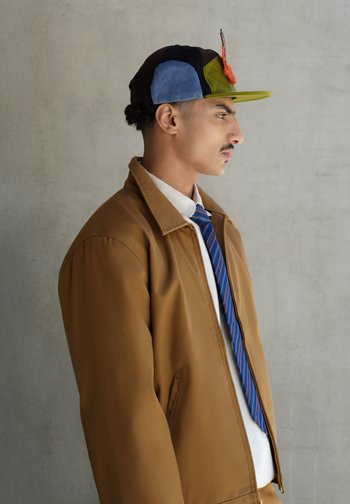 Jaqueta bomber castanha, camisa branca e gravata às riscas azul. Boné multicolorido com apliques e detalhe de pena. Textura suave, corte estruturado.