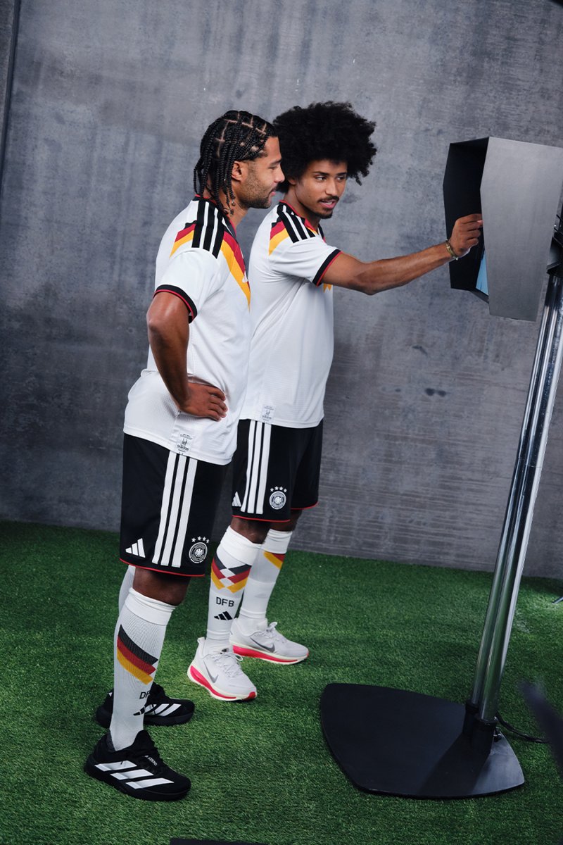 Zwei Männer in weißen und schwarzen Fußballtrikots mit Deutschlandflaggen-Design. Einer steht mit den Händen in der Hüfte, während der andere mit einem Gerät interagiert.