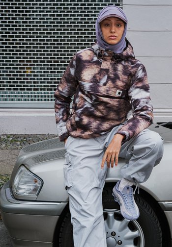 Person som bär en lavendel hijab, mönstrad jacka och ljusgrå cargobyxor, lutar sig mot huven på en silverfärgad bil som är parkerad på en stadsgata.